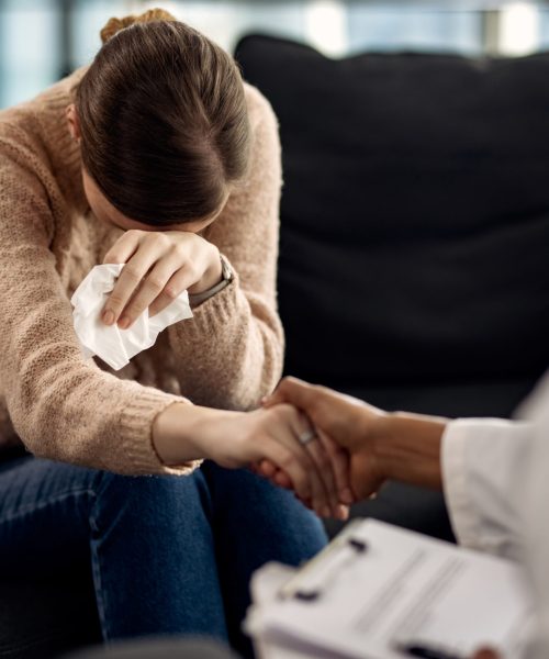 Sad woman crying while holding hands with her psychotherapist at doctor's office.