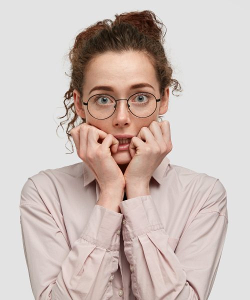 Unhappy nervous young female with worried expression, bites finger nails, looks anxiously directly at camera, wears spectacles, dressed in fashionable clothes, stands against white background.